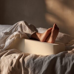 A peaceful, high-end bedroom scene showing a person’s legs resting comfortably on a specialized ergonomic wedge pillow. The bedding is high-quality white linen, and the lighting is soft, warm, and evening-toned. Focus on the elevation of the legs.